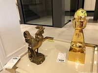 Photo of bronze rooster statue and gold-colored Asian metal art object together on a table.