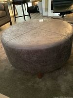 Front-left view of round ottoman showing leather upholstery and wooden leg