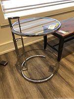 Side table in room next to wooden bench showing chrome frame and round clear glass top.