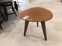 Front angle view of triangular wood side table with three tapered legs on a tiled floor.