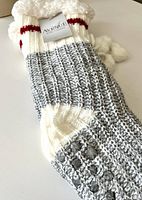 Photo of a single knit sock showing the gray and white ribbed pattern, red stripe near cuff, and white pom-poms, along with visible silicone grip dots on sole.