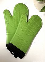 Pair of green silicone oven mitts, textured with diamond-pattern grip, shown lying side by side with black fabric lining at cuff.