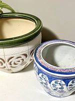Close-up of the tops of the two plant cover pots showing the green rimmed cream pot with rose carvings and the blue pot with floral patterns