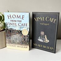 Front view of the two hardcover books by Stuart McLean titled 'Home from the Vinyl Cafe' and 'Vinyl Cafe Unplugged' standing upright side by side on a table next to a plant.