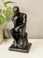 Front view of the black resin 'Thinking Man' statue showing the muscular figure seated with hand on chin on a square base.