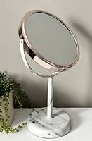 View of round double-sided vanity mirror with rose gold edging and white marble patterned stand on a light surface, neutral wall background.