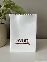 Front view of the white ceramic vase shaped like a crumpled paper bag with 'Avon' logo in black and red on one side.