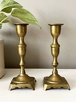 Front view of the two vintage brass candle holders side by side on a surface with a plant nearby.