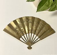 Front view of brass decorative fan wall hanging showing embossed floral and plant details and overall fan shape.