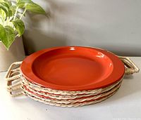 Stack of four bright orange ceramic plates in individual wicker and metal holders with woven handles, placed on a white surface near a green plant.