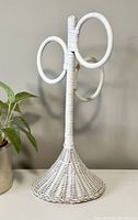 Full view of a white wicker hand towel rack standing upright against a wall with a plant pot nearby.