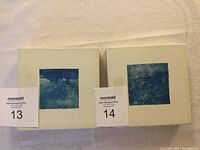 Two small square mounted artworks with abstract blue ocean hues, labeled with lot numbers 13 and 14, showing surface condition and mounting