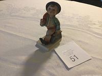 Photo of a porcelain Hummel figurine showing a boy with an umbrella standing on a white base with a lot number card beside it.