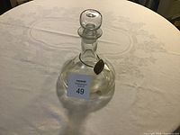 Clear glass sherry decanter with round stopper and brass label on neck, on white patterned tablecloth.