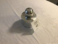 Closed view of small silver cylindrical container with polished surface and spherical knob lid, placed on a light surface.