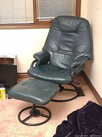 Swiveling dark green leather recliner chair with matching ottoman in corner near windows showing chair and ottoman with cushioned leather and metal bases.