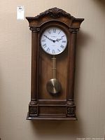 Front view of entire wooden wall clock showing the decorative top, clock face with Roman numerals and brass pendulum beneath glass.