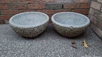 Two round concrete planters side by side showing full exterior and pebbled texture, placed on concrete floor against brick wall.