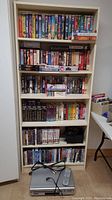 Photo showing a shelf filled with approximately 120 VHS tapes and DVD movies, with the JVC VHS/DVD player and remote placed on the floor in front of the shelf.