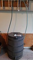 Full view of the dark brown plastic rain barrel standing in a garage, showing connected hose and vents.