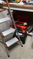 Aluminum 3-step stepladder with orange accents next to black plastic stepstool and stack of tools in the basement