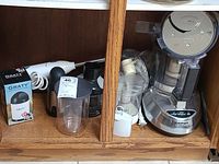 View of multiple small kitchen appliances including Cuisinart food processor, KitchenAid immersion blender, Black & Decker food processor, cheese graters and accessories inside cabinet
