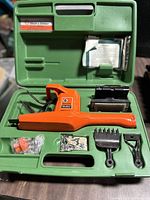 Photo showing the open green plastic case with an orange Black & Decker electric tool and various attachments including blades, rakes, hardware pieces, and manual.