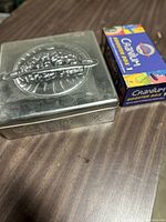 Metal Cranium game box with visible wear along edges and embossed logo on lid, placed next to colorful Cranium Booster Box 1