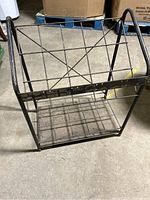 Front view of a black metal rack with two shelves and geometric side supports. Shows rusty patina on metal surfaces.