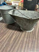 Photo showing two galvanized metal buckets on a wooden surface, the coal bucket with a wire handle is slightly smaller and tapered, the larger one is more cylindrical.