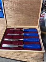 Four blue-handled wood chisels inside a wooden box with red felt lining.