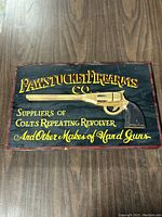 Front view of wooden sign with painted text and a three-dimensional revolver motif in the center on a black background.