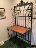Front angled view of a black powder coated metal bakers rack with arborite wood grain base and decorative curled metal sides and top crest.