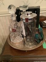 Crystal cut decanter with stopper, cocktail shaker, tartan bottle cosy, and silver plated tray on wooden table