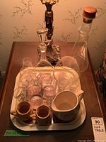 Famous Grouse barware items arranged on a wooden side table featuring a tray holding multiple glasses, two small mustard pots, a ceramic pitcher, and two glass decanters.
