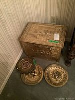 Photo showing brass fireside wood storage box and three brass plates on carpeted floor near wall.