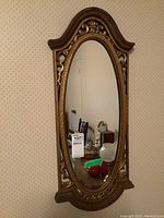 Full view of the vintage oval wall mirror showing the carved gilded wood frame with intricate scroll and leaf details, mounted on a patterned wallpapered wall.