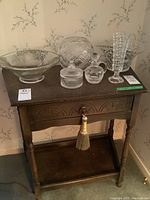 Front and top view of oak occasional table showing single drawer with decorative carving and brass pull with tassel, holding various crystal and glass items (not included).
