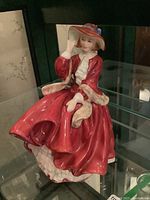 Front and side view of the Royal Doulton figurine showing detailed red dress, white ruffled underskirt, and red hat with blue ribbon.