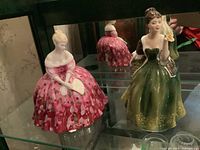 Two Royal Doulton figurines displayed in a glass cabinet, one in a pink floral dress holding a fan, the other in a green and brown dress holding a flower.