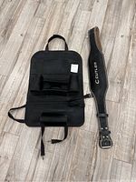 Black fabric car seat back organizer laid flat on wooden floor along with a black belt labeled 'GUTTLER'.