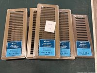 Four brown louvered floor registers and one brown floor register with solid slats shown side by side.