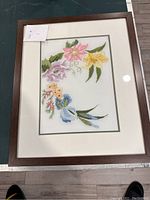 Framed flower embroidery artwork showing colorful flowers with pink, purple, yellow, blue, and green leaves inside a wooden frame with white matte border. Measuring approximately 44 cm by 36 cm.