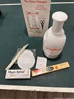 Photo showing The Euro Chopper with white plastic body and clear container section, boxed Euro Chopper packaging, Magic Spiral box, a white rice paddle, circular lid accessory, and a pair of black-handled scissors.