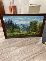Front view of the large framed oil painting showing a forest landscape with mountains and cloudy sky.