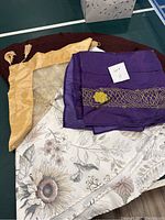 Four table cloths folded and stacked: purple with gold embroidery, yellow satin with tassels, floral neutral print, and gold embroidered semi-sheer.
