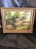 Frontal view of framed oil painting showing forested landscape with large rocks and trees, capturing the entire frame and artwork, highlighting the condition of frame and artwork.