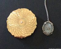 Top view of the gold-tone woven textured Stratton compact next to the oval sterling silver and camphor glass pendant necklace with the Eastern Star symbol.