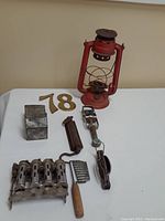Vintage lot items displayed on white surface included red metal lantern, brass numbers 7 and 8, small coin holder box, match holder with fork tool, vintage kitchen/mechanical tool with wooden handle and connected stove burner covers.