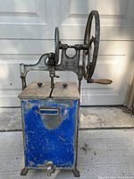 Front side view showing the blue metal body, wooden top lids, metal churning mechanism with wheel handle and cogs.